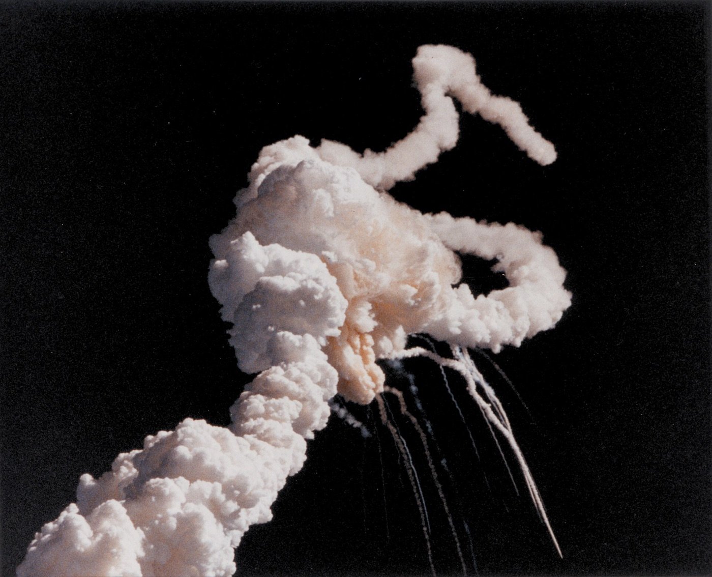 A white smoke plume spiralling against a dark sky with booster trails falling below