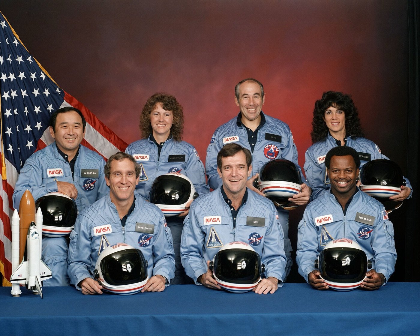 The seven STS-51-L crew members in blue flight suits holding helmets in front of a flag