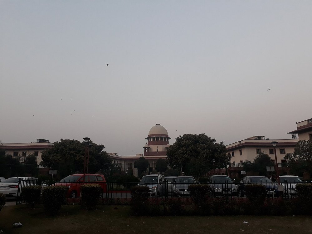 The domed Supreme Court of India building seen from across a railing and parked cars at dusk