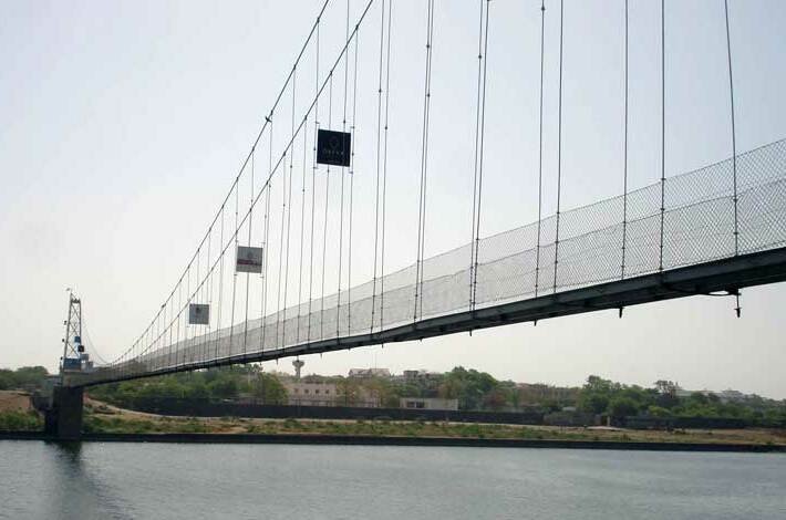 Side view of the Morbi suspension bridge with vertical hangers, a mesh-sided deck and a tower over the Machchhu River