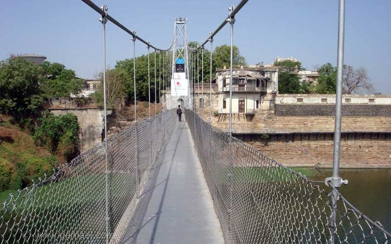 A narrow pedestrian walkway with mesh sides on the Morbi suspension bridge, with a tower and old buildings ahead