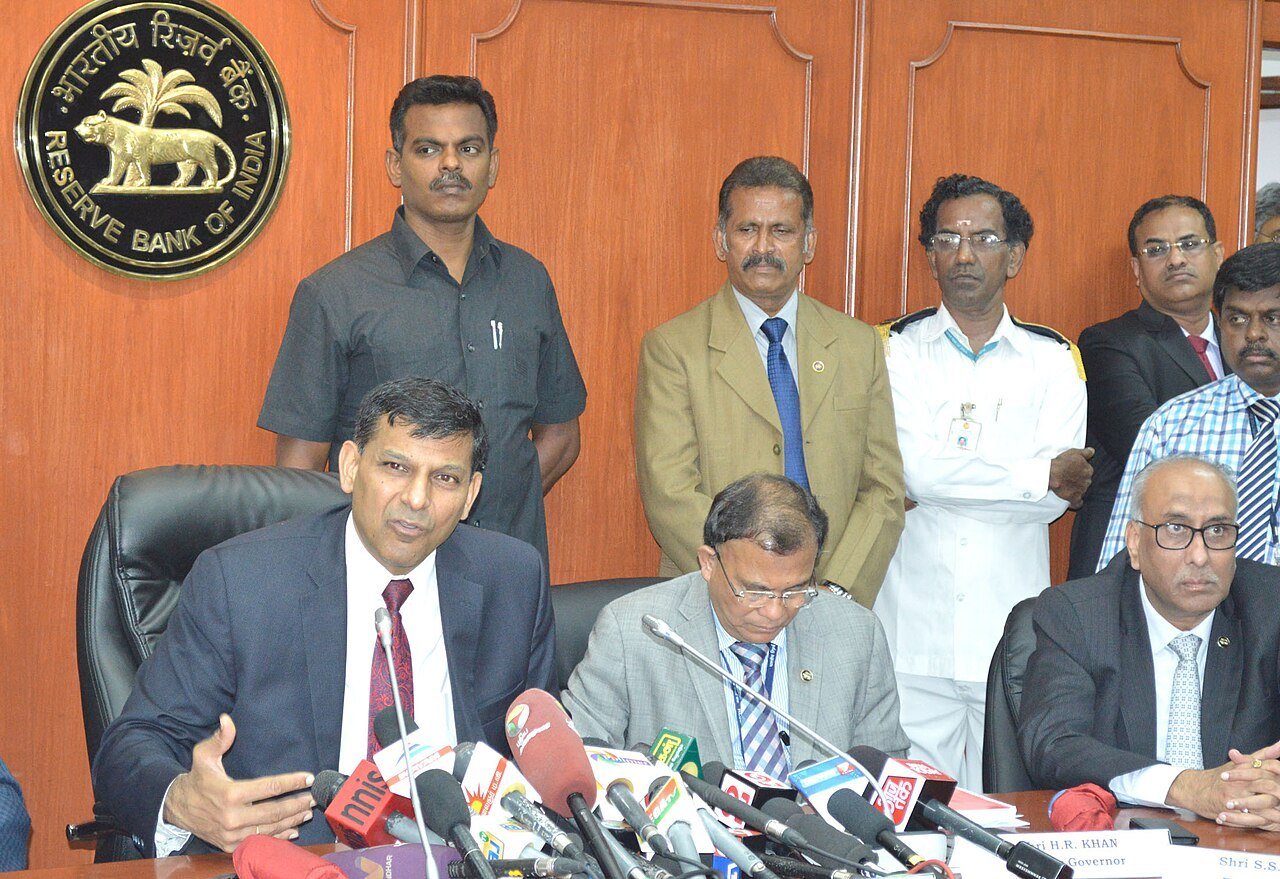 RBI Governor Raghuram Rajan seated at a table with microphones in front of the Reserve Bank of India emblem, flanked by officials