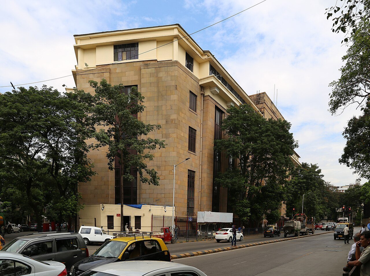 A multi-storey beige Reserve Bank of India office building on a Mumbai street corner with trees and a taxi in front