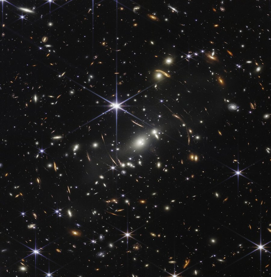A dense field of thousands of galaxies, some stretched into arcs by gravitational lensing around the cluster SMACS 0723, against black space with a few bright foreground stars