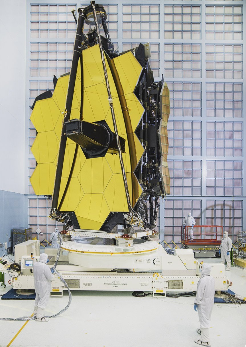 The gold-coated primary mirror of the James Webb Space Telescope in a cleanroom with one wing folded back, with technicians in white suits below