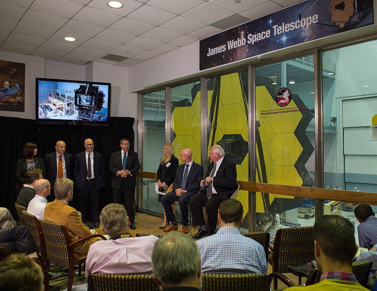 Officials and journalists seated in front of a glass wall behind which the gold hexagonal mirror segments of the James Webb Space Telescope are visible, with a Webb sign above