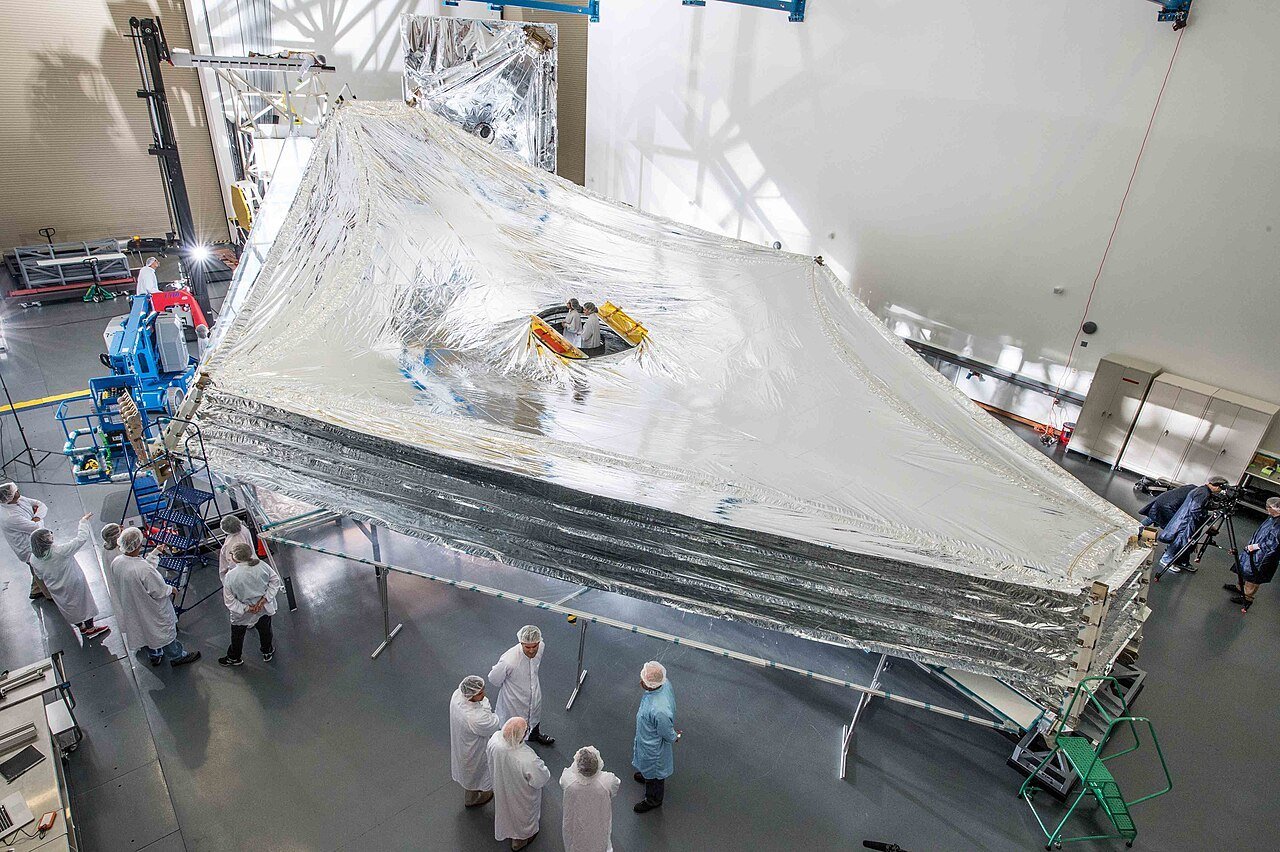 The silver five-layer sunshield of the James Webb Space Telescope spread out in a large cleanroom while technicians in white suits inspect it