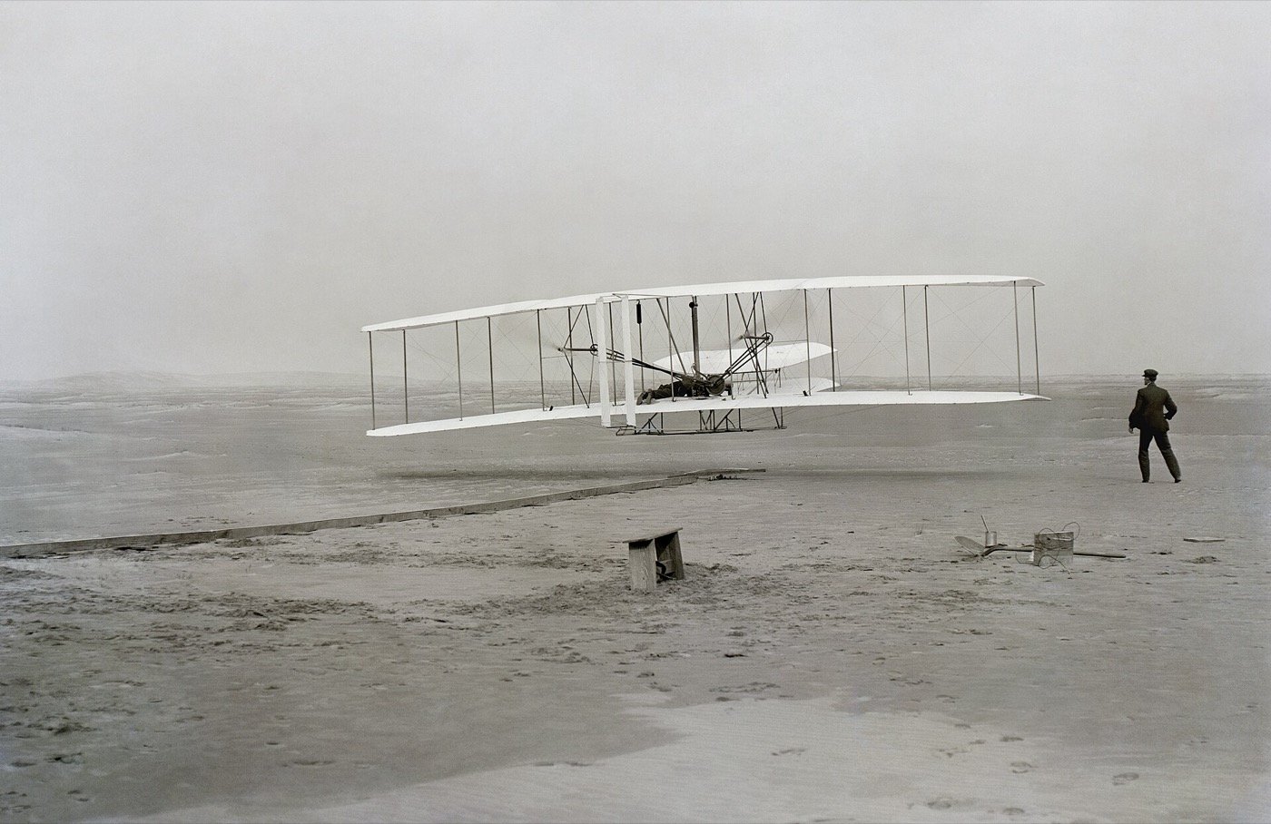 The 1903 Wright Flyer a few feet above the sand at Kitty Hawk with a man watching on the right